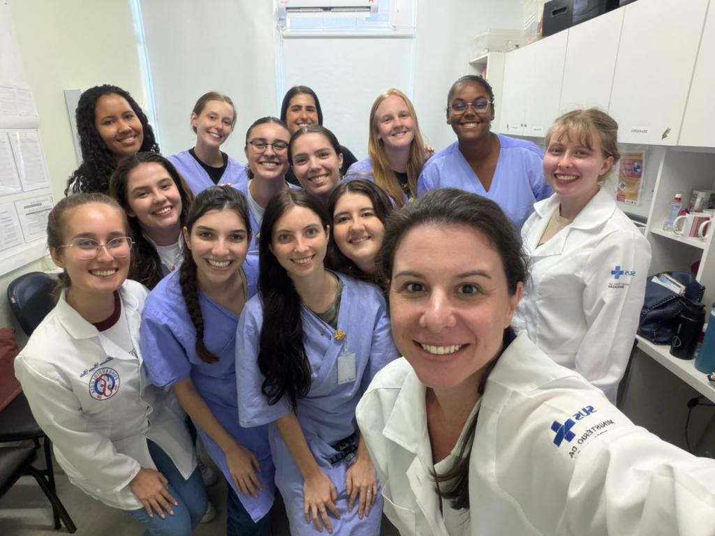Foto selfie, horizontal, colorida com um grupo de 13 profissionais de saúde sorrindo. A maioria veste uniformes azuis ou jalecos brancos com o logotipo do SUS e Ministério da Saúde, em um ambiente de clínica.