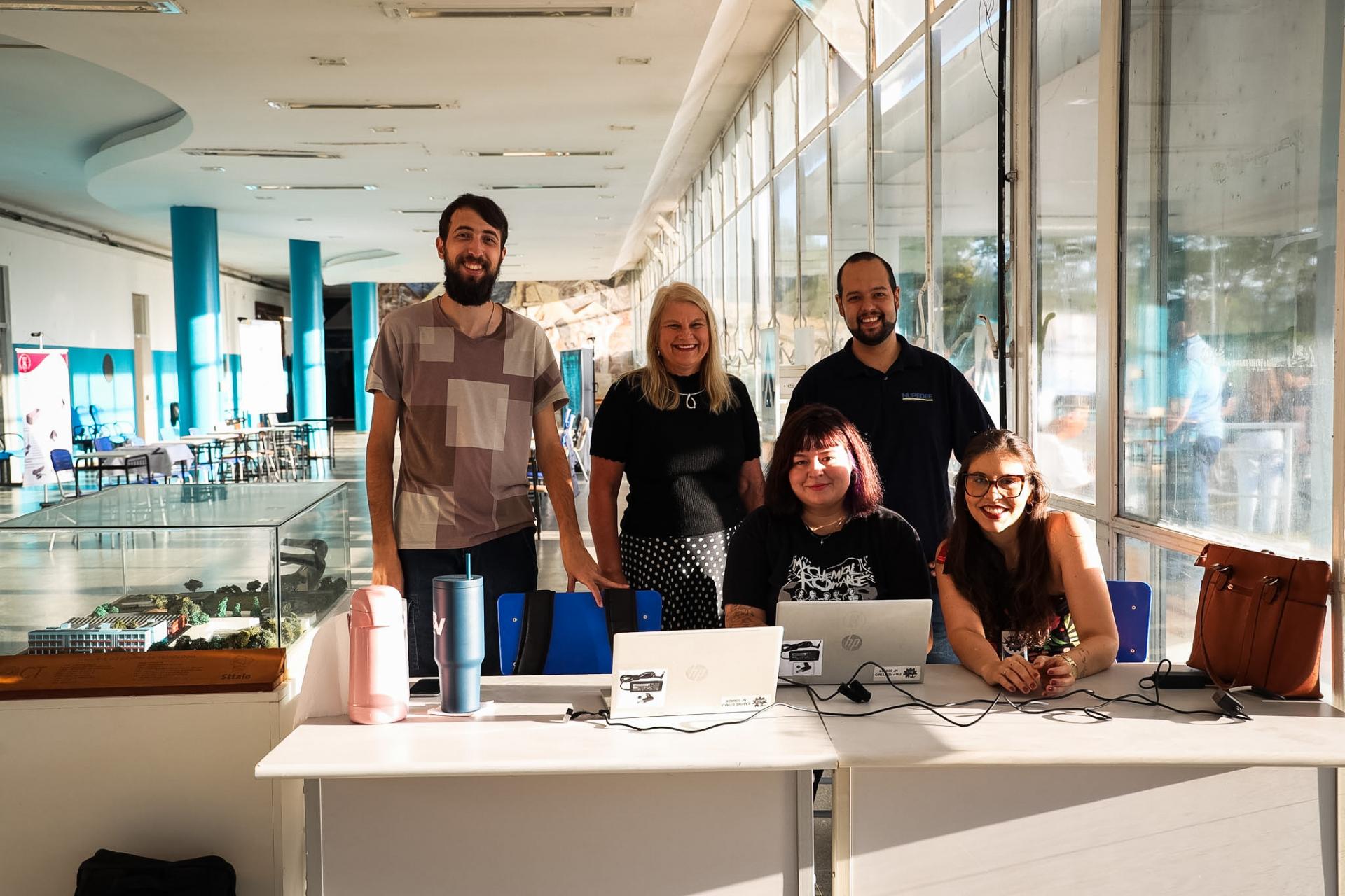 Foto colorida horizontal de cinco pessoas no hall do Centro de Tecnologias. Duas melhores estão sentadas atrás de um balcão de atendimento e as outras três estão em pé atrás delas.