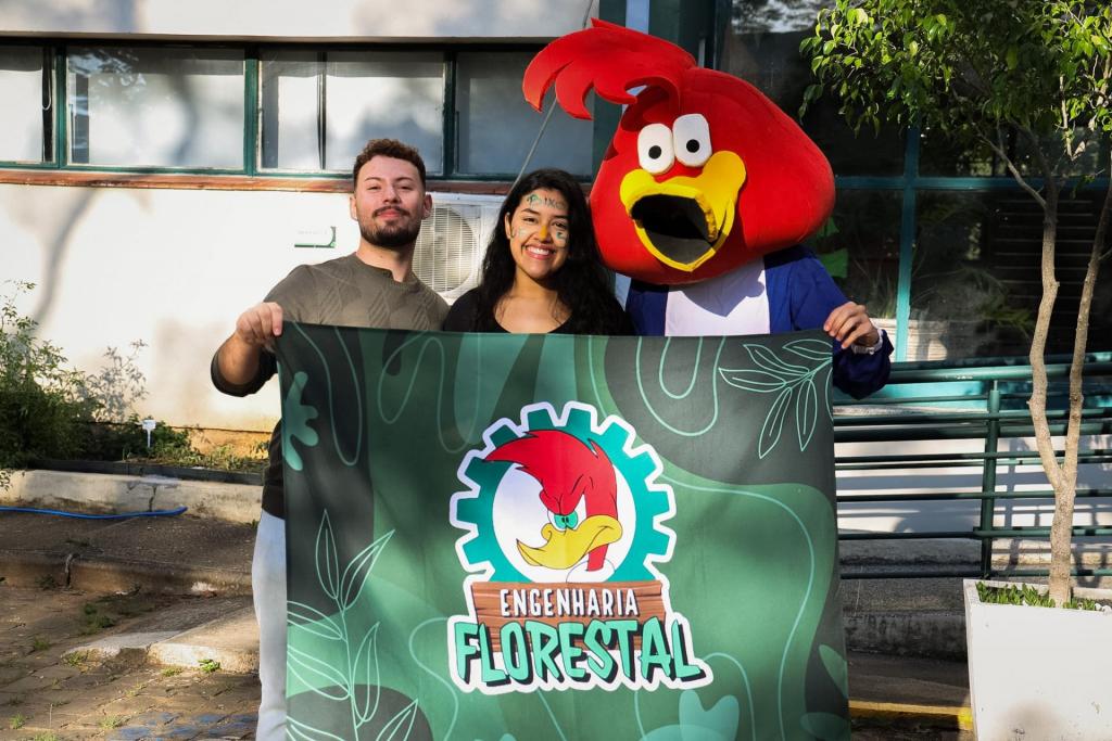 Foto colorida horizontal de três pessoas segurando uma faixa do curso de Engenharia Florestal. Da esquerda para a direita, um homem jovem de barba, uma mulher jovem de cabelo escuro e longo, e uma pessoa fantasiada de Pica-Pau