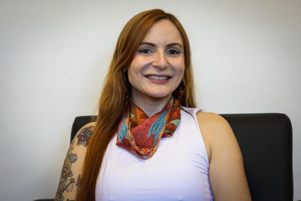 Foto horizontal colorida de uma mulher branca, adulta, com cabelos longos e lisos na cor ruivo. Ela está sentada em uma cadeira preta, em frente a uma parede clara e lisa. Veste uma blusa sem mangas de cor branca e um lenço colorido no pescoço. No braço, há várias tatuagens coloridas visíveis. A imagem tem enquadramento médio, focado do peito para cima.