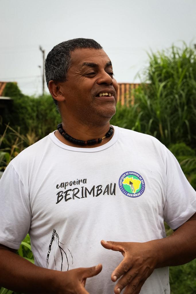 Foto vertical colorida de um homem negro, de meia-idade e sorrindo. A imagem o mostra do peito para cima e olhando para o lado direito. Ele tem cabelos curtos e grisalhos, usa uma camiseta branca com a inscrição “Capoeira Berimbau” e um emblema colorido no peito. No pescoço, utiliza um colar de contas escuras. Suas mãos aparecem à frente do corpo, como se estivesse gesticulando enquanto fala. Ao fundo, desfocado, há vegetação verde alta. O céu aparece nublado acima da paisagem.