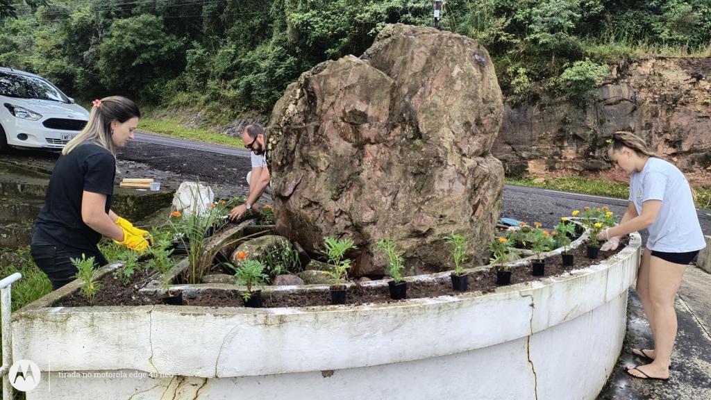 Um homem e duas mulheres plantam flores em um canteiro localizado em torno a uma grande rocha. O canteiro fica à beira de uma estrada asfaltada, uma ladeira bastante inclinada aos pés de um morro. À esquerda, está estacionado um carro branco.