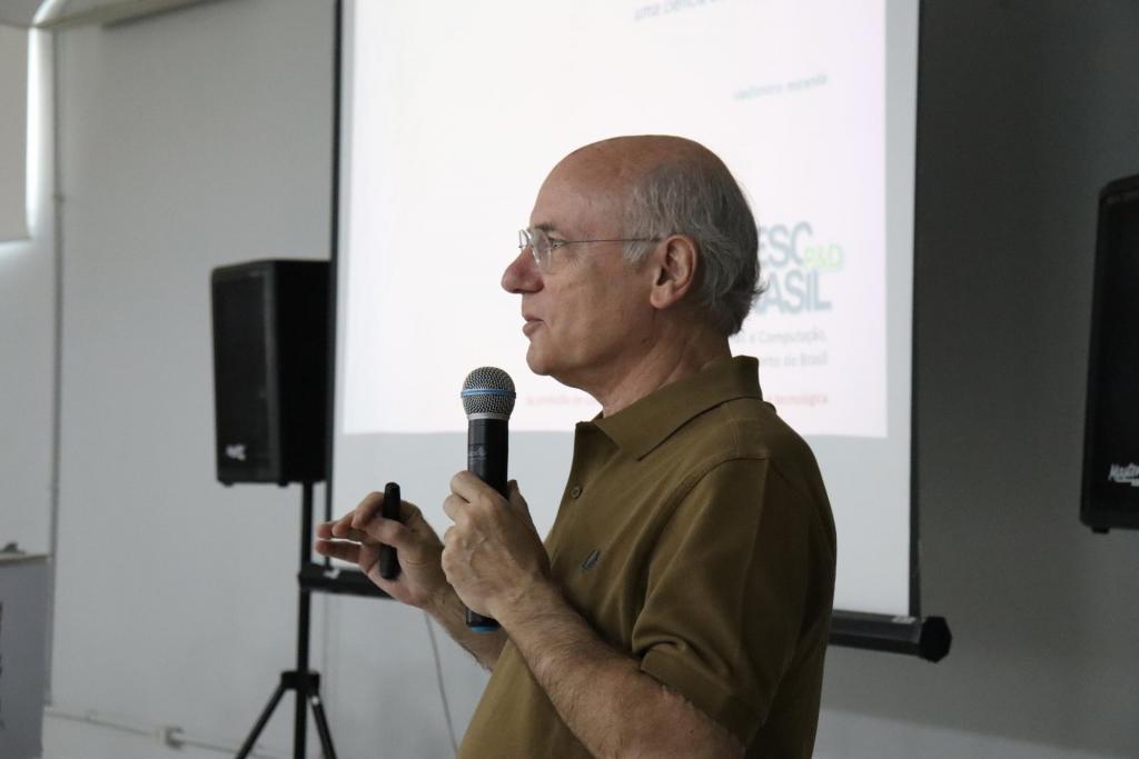 Foto horizontal e colorida do perfil de Vladimiro Miranda. Ele segura um microfone enquanto fala ao quórum presente na palestra