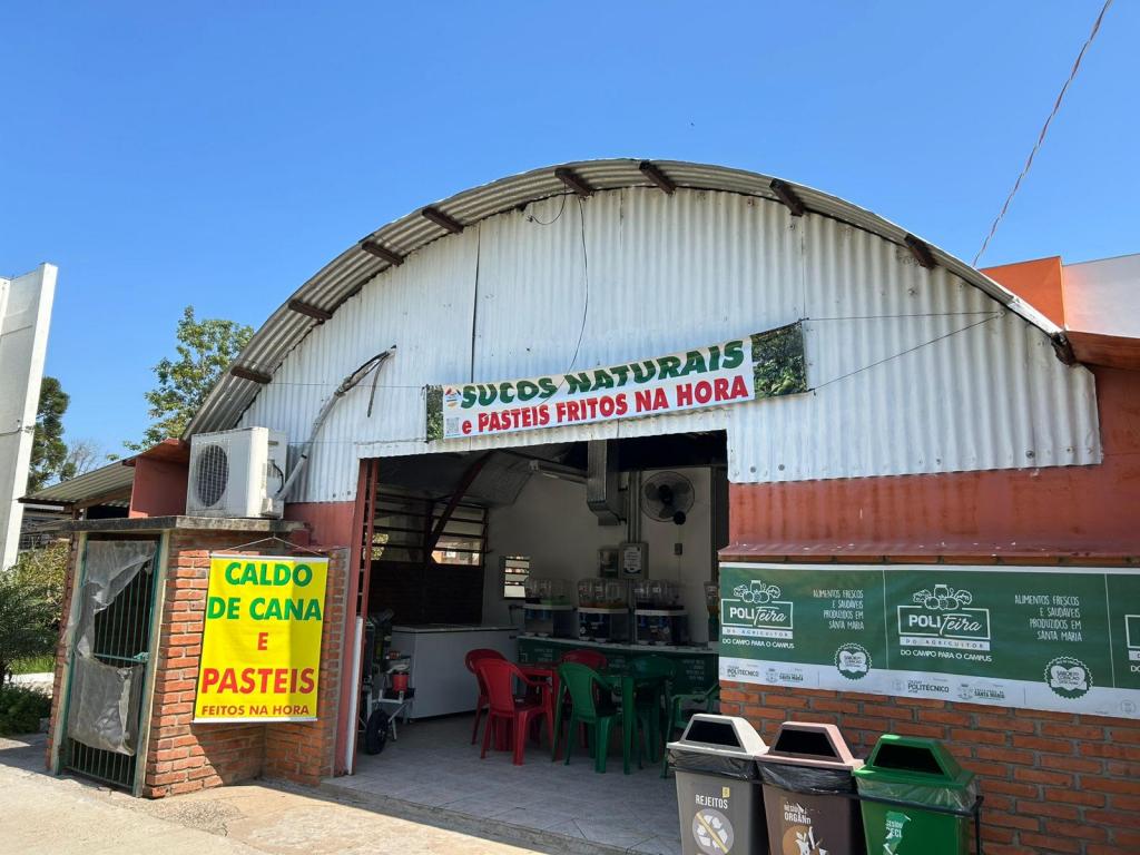 A foto mostra uma edificação rústica (semelhante a um galpão) com base de tijolos e telhado de placas de metal. Na fachada, estão afixados cartazes que anunciam os produtos vendidos pela PoliFeira.