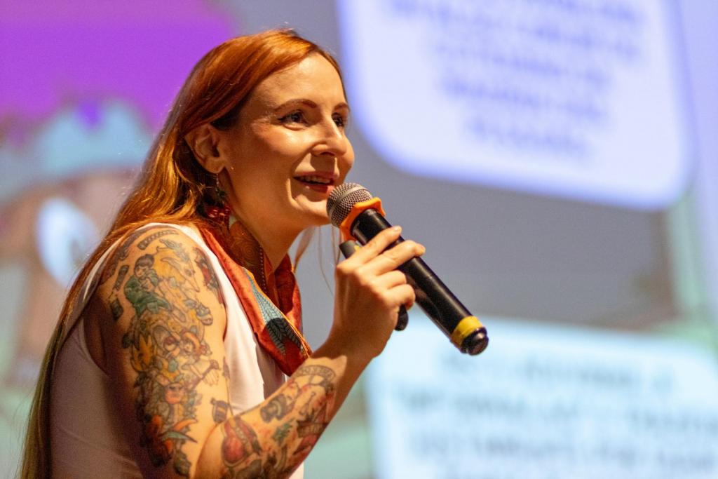 Foto colorida horizontal de mulher ruiva com blusa branca e lenço vermelho. Ela está de lado e segura um microfone. Atrás dela uma projeção colorida. A foto mostra a mulher de lado e do peito para cima.