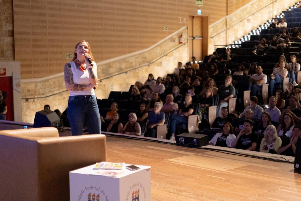 Foto colorida horizontal de mulher em pé no palco do centro de convenções. Atrás dela a plateia