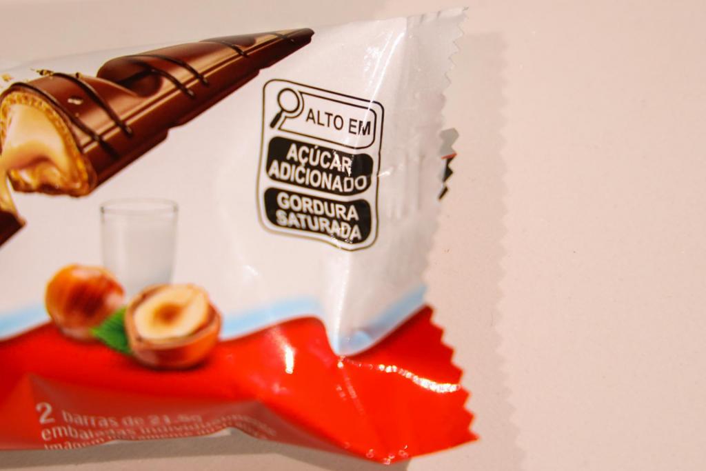 Fotografia horizontal, colorida, em close de uma embalagem de suco industrializado de laranja com fundo branco. No centro há a ilustração de uma fatia de laranja estilizada com elementos gráficos coloridos. A embalagem traz mensagens promocionais como “fonte de vitamina A e zinco” e “contém 10% de fruta”, além da indicação de volume de 200 ml.