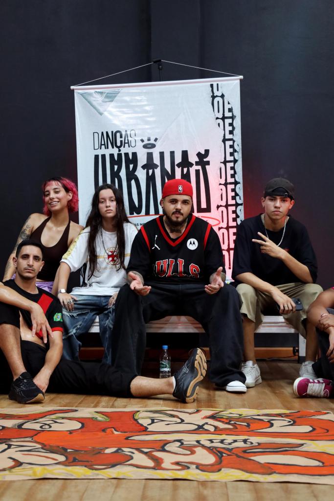 Foto na vertical e colorida que mostra, ao centro, Mauricio com alguns dos dançarinos presentes no evento, todos sentados. No chão, um cartaz colorido com traços em laranja. Ao fundo, o mesmo cartaz exibindo o nome do projeto e a logo e atrás disso, uma das paredes pretas do estúdio