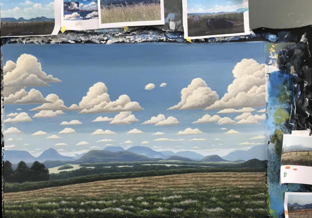 Foto horizontal. Mostra um quadro pintado, representando um campo com algumas árvores e montanhas ao fundo, sob um céu repleto de nuvens. Em volta do quadro, fixadas em uma parede colorida com diversas cores, estão fotos que possivelmente serviram de inspiração para a pintura.