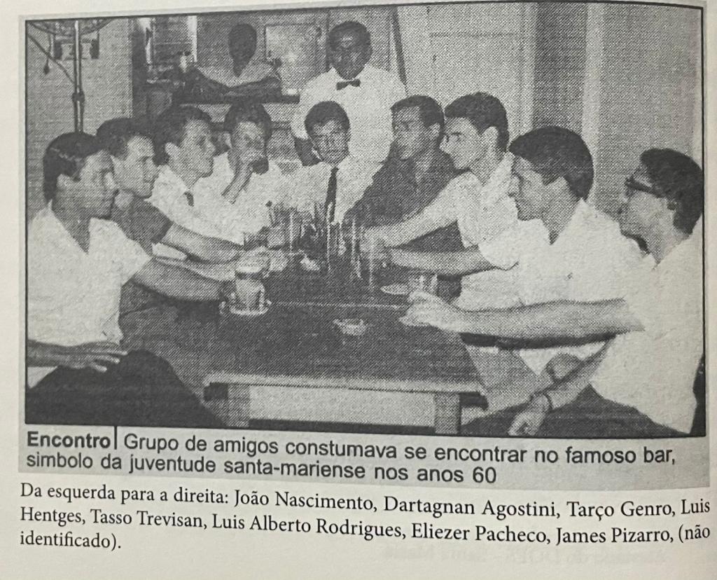 Foto em tons de cinza. semelhante a um recorte de jornal, com dez homens, sendo oito sentados à mesa. Todos estão com camisas e têm cabelos curtos escuros ou claros. Alguns usam óculos. Os outros dois homens são o garçon e um home no balcão ao fundo.