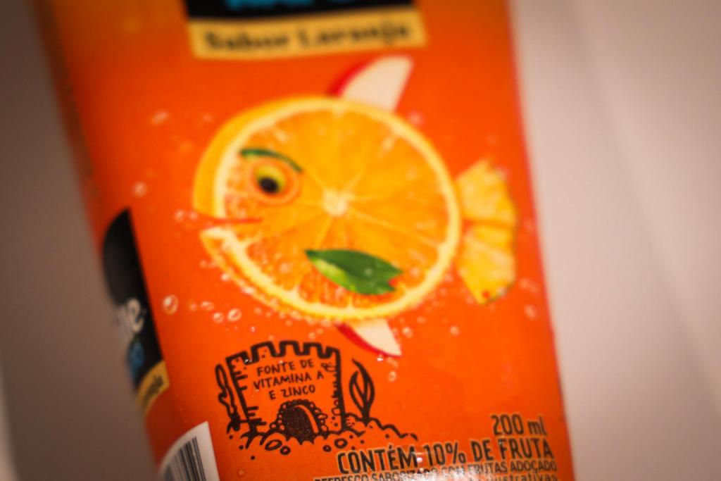 otografia horizontal, colorida, em close de uma embalagem de suco industrializado de laranja com fundo branco. No centro há a ilustração de uma fatia de laranja estilizada com elementos gráficos coloridos. A embalagem traz mensagens promocionais como “fonte de vitamina A e zinco” e “contém 10% de fruta”, além da indicação de volume de 200 ml.