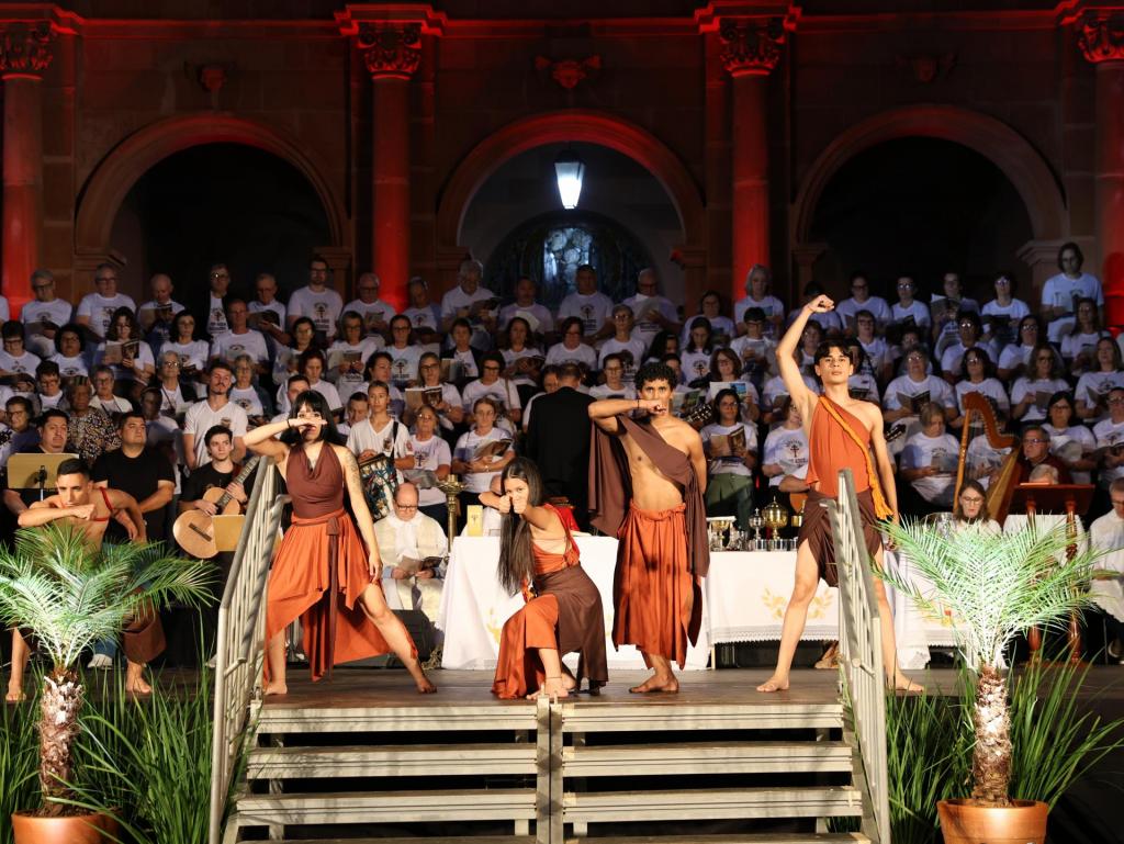 Em frente à catedral, está posicionado um coral formado por dezenas de pessoas vestidas com camisetas brancas. Em frente ao coral, sobre um estrado, quatro jovens bailarinos (dois meninos e duas meninas) representam índios no espetáculo.