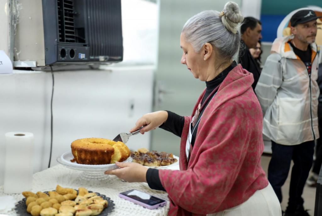No centro da foto, está uma mulher branca de cabelos brancos, que veste uma blusa de cor rosa avermelhada, sobre uma camisa preta de gola rolê. Ela aparece de perfil, em frente a uma mesa com petiscos variados. Com uma espátula na mão, ela corta pedaços de bolo.