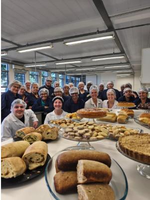 Foto colorida vertical de grupo de pessoas uniformizadas com aventais brancos e azuis, bem como todas brancas. Todos estão atrás de uma mesa com alimentos dispostos, como pães, pães recheados salgados e bolos doces