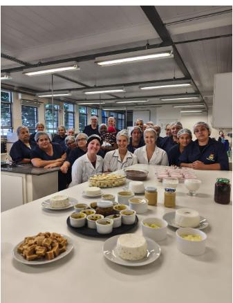 Foto colorida vertical de um grupo de pessoas sentadas em bancos e atrás de bancadas. O grupo veste aventais brancos e azuis escuros com todas brancas. Todos estão sentados atrás de uma mesa com vários alimentos dispostos, como queijo, biscoitos e molhos