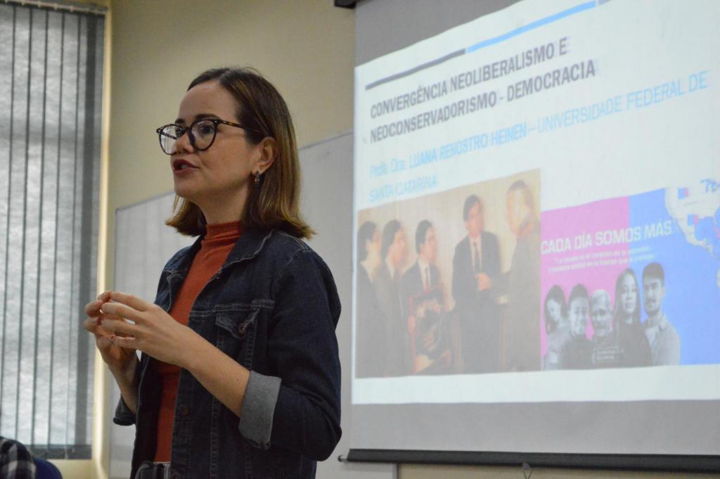 Foto colorida horizontal da palestrante em pé, em imagem da cintura para cima e da esquerda para a direita. Luana veste óculos, uma blusa cor telha e um casaco jeans. Atrás dela, a projeção de um slide
