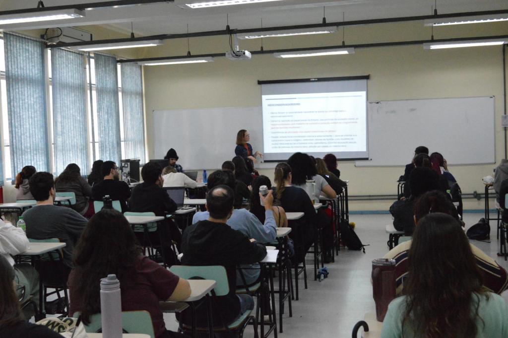 Foto colorida horizontal de sala de aula lotada. Ao fundo, a palestrante Laura à frente da projeção. Em fila, sentados, os estudantes do curso de Relações Internacionais.