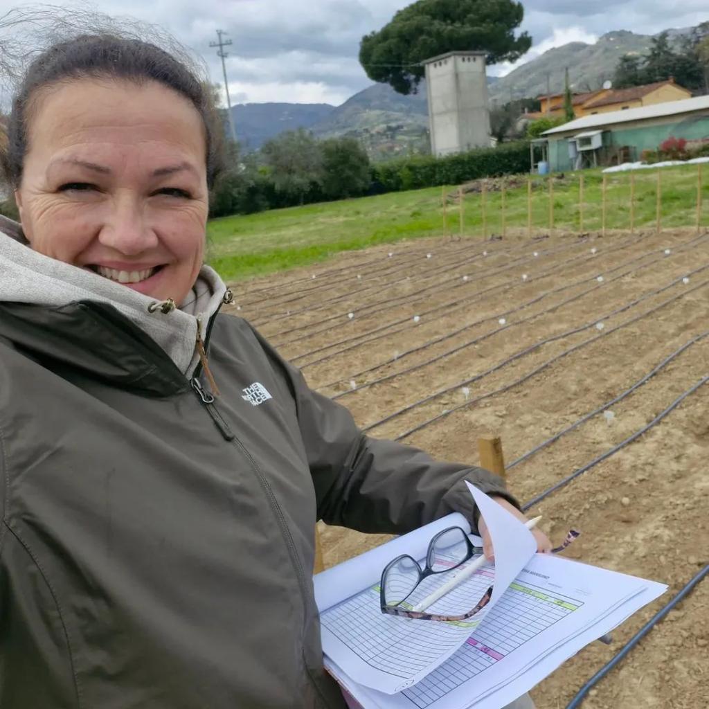 Foto colorida vertical em formato selfie de mulher adulta branca de cabelo escuro e preso. Ela veste um casaco escuro e segura uma prancheta com uma folha euma caneta. A imagem mostra ela no campo, à frente de uma plantação.