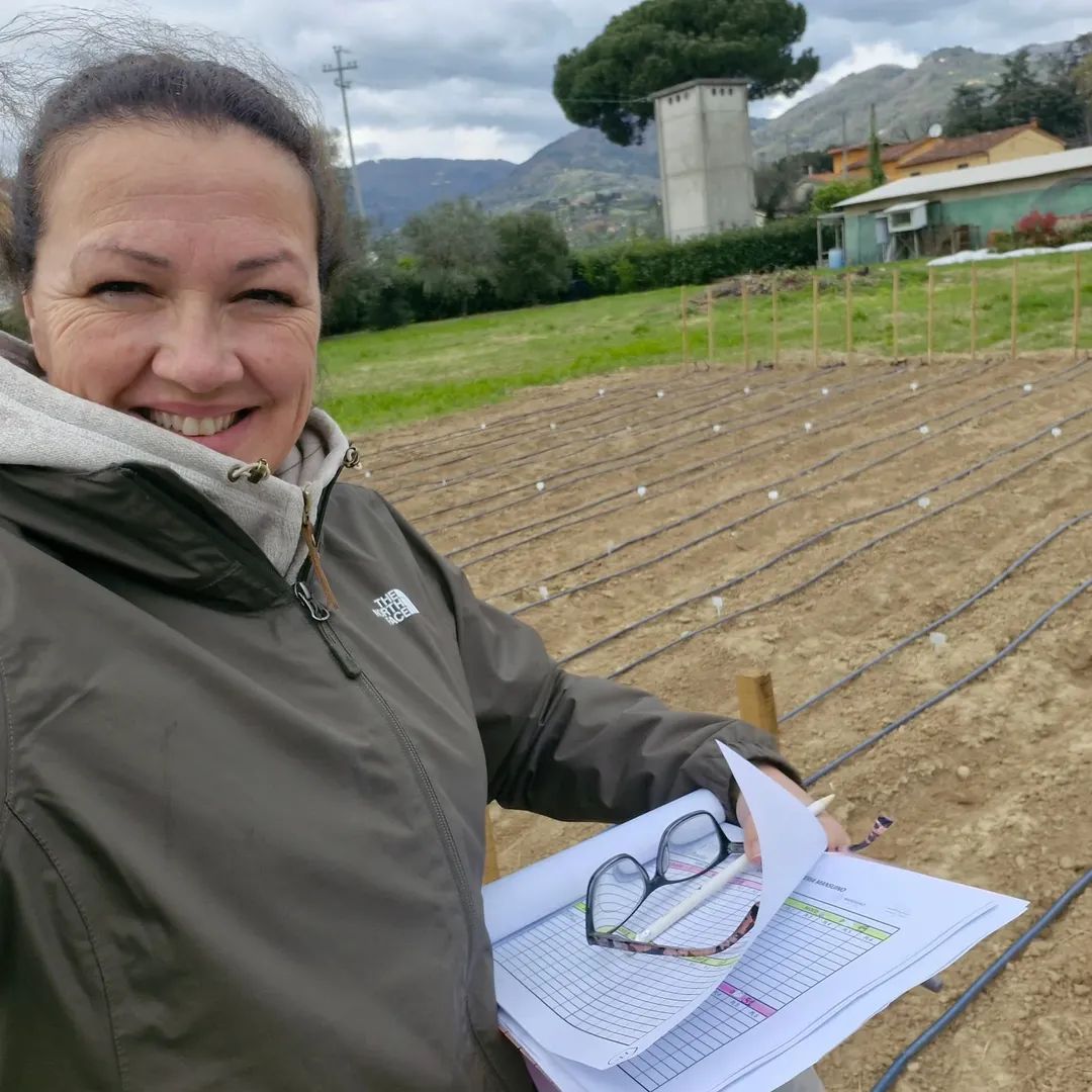 Foto colorida vertical em formato selfie de mulher adulta branca de cabelo escuro e preso. Ela veste um casaco escuro e segura uma prancheta com uma folha euma caneta. A imagem mostra ela no campo, à frente de uma plantação.