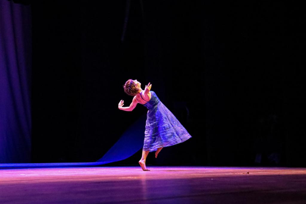 Foto colorida horizontal de mulher em uma palco amplo dançando.Ela usa um vestido azulado e está com o corpo posicionado à direita.