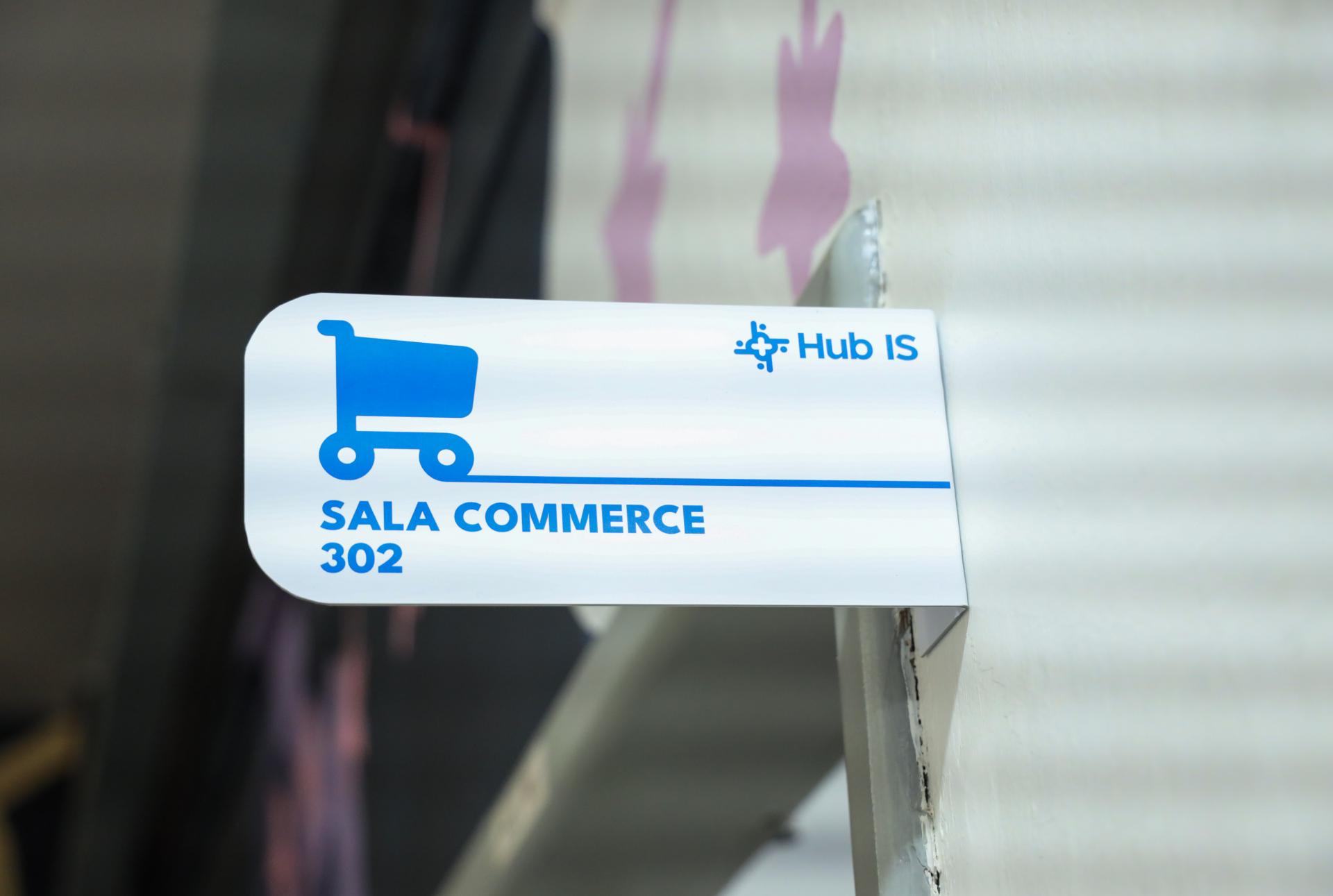 A foto mostra uma placa com a inscrição “Hub Is / Sala Commerce / 302”. Na placa, destaca-se também o desenho em azul de um carrinho de supermercado.
