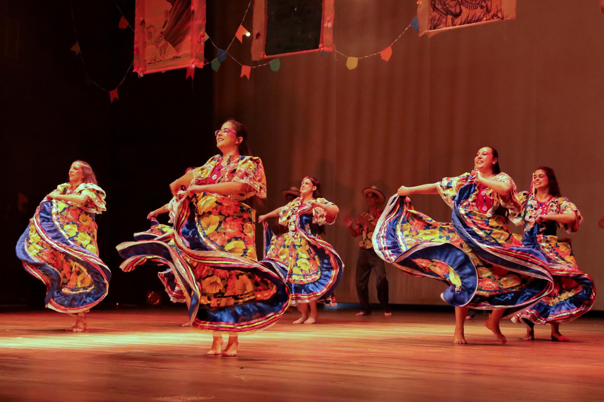 Foto colorida horizontal de cinco mulheres com vestidos rodados, coloridos com temáticas regionais. As bailarinas dançam no palco do Centro de Convenções e agitam seus festidos coloridos.