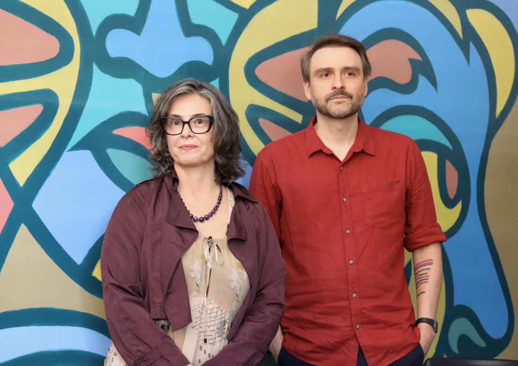 Uma mulher e um homem posam para a foto, tendo como fundo uma parede multicolorida. A mulher é branca e grisalha, usa óculos, veste uma jaqueta grená e usa no pescoço um colar da mesma cor; sob a jaqueta, usa um vestido bege. O homem é branco e grisalho, tem uma barba curta e veste uma camisa vermelha.