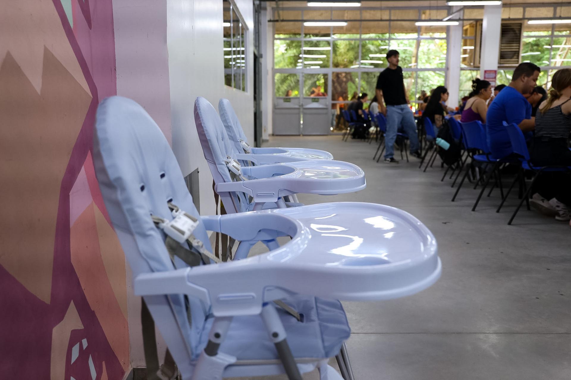 Foto horizontal colorida. Em destaque, a esquerda da imagem, três cadeiras de alimentação infantil. Ao fundo, mesas e cadeiras do Restaurante Universitário.