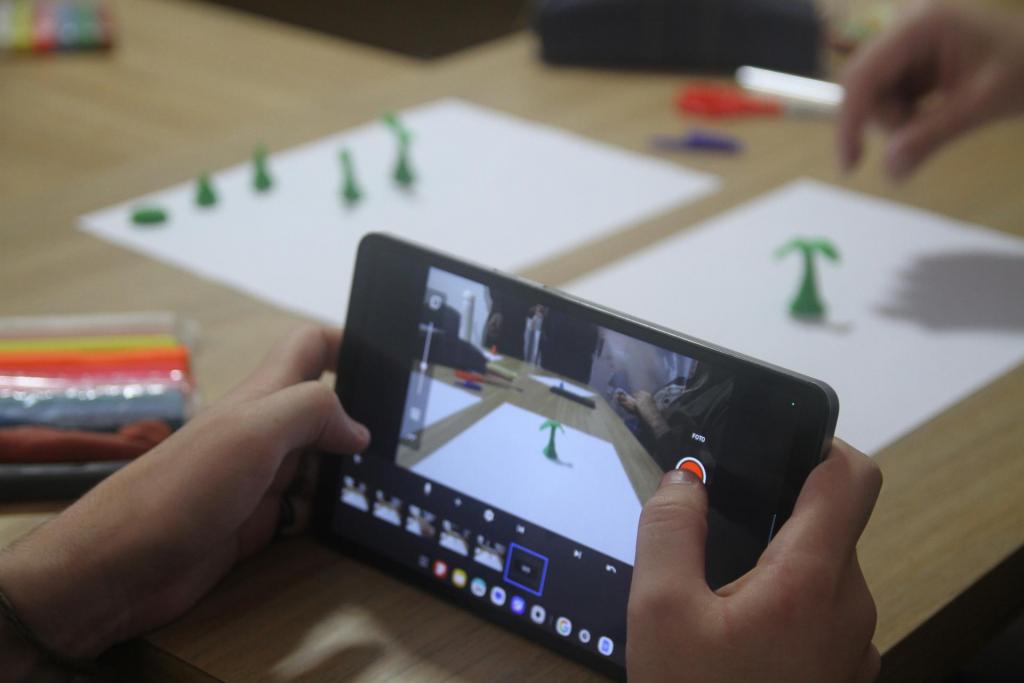 Foto colorida, em formato horizontal, mostra ao centro duas mãos segurando um tablet com a câmera aberta, utilizado para gravar um vídeo em stop-motion com massinha de modelar. Sobre a mesa, há uma folha branca e, sobre ela, uma massinha verde modelada em forma de planta, imagem registrada na tela do aparelho. Ao fundo, em desfoque, aparecem outras peças de massinha já modeladas para compor o restante da animação, além de porções de massinha de diferentes cores ainda não utilizadas