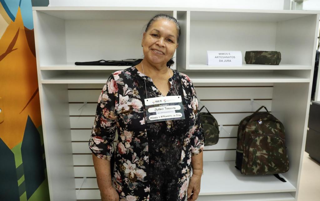 A foto mostra uma mulher morena com cabelos grisalhos, que vete uma camisa com estampa flores e fundo escuro. Ela está postada à frente de uma estante na qual estão em exposição artigos com decoração de camuflagem militar.