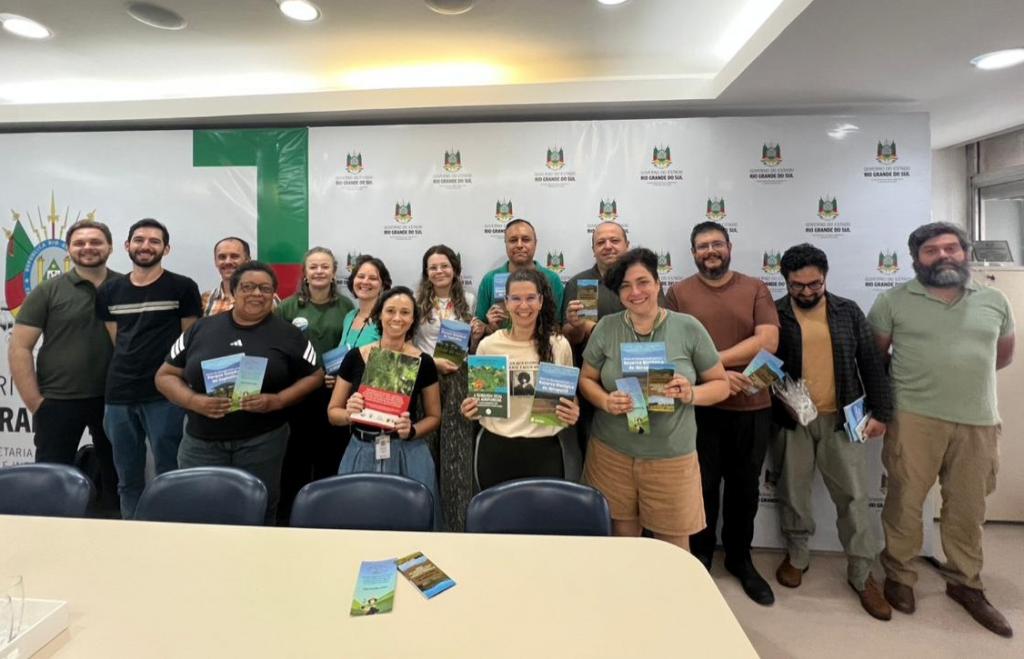 Em pé e atrás de uma mesa com cadeiras, 15 pessoas posam para a foto, segurando livros e fôlderes.