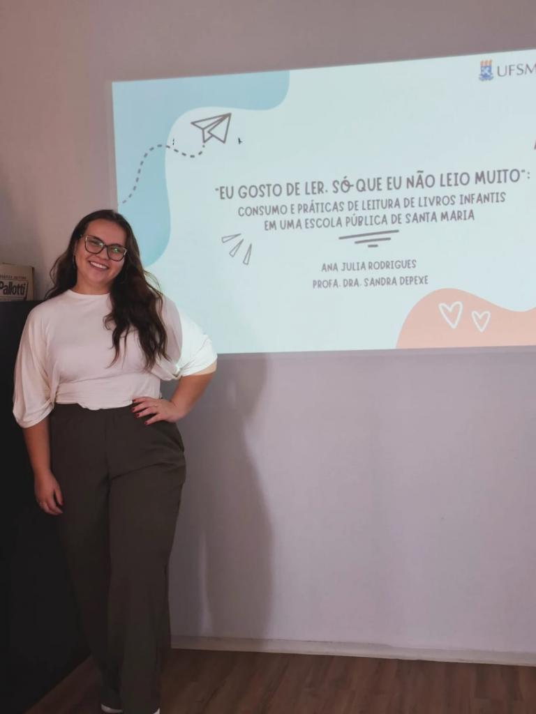 Foto colorida vertical de mulher jovem em pé. Ela usa óculos, tem cabelos claro longo, usa blusa branca e calça escura. Atrás dela, a projeção de um slide com o nome da pesquisa “‘Eu Gosto de Ler, Só Que Eu Não Leio Muito’: Consumo e Práticas de Leitura de Livros Infantis em uma Escola Pública de Santa Maria”
