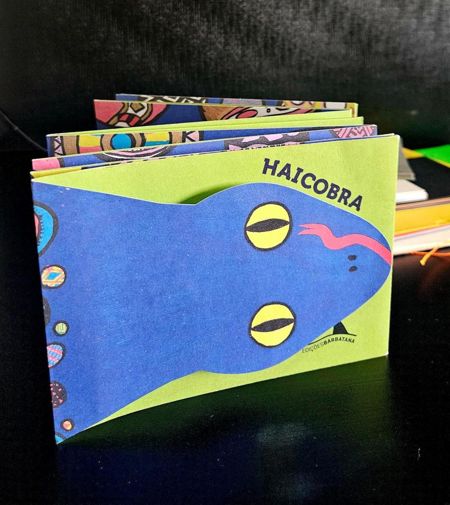 Foto colorida vertical de livro infantil com desenho de cobra azul sobre um fundo amarelo. A cabeça da cobra acompanha o formato da capa da obra, que tem dois olhos e um lingua.