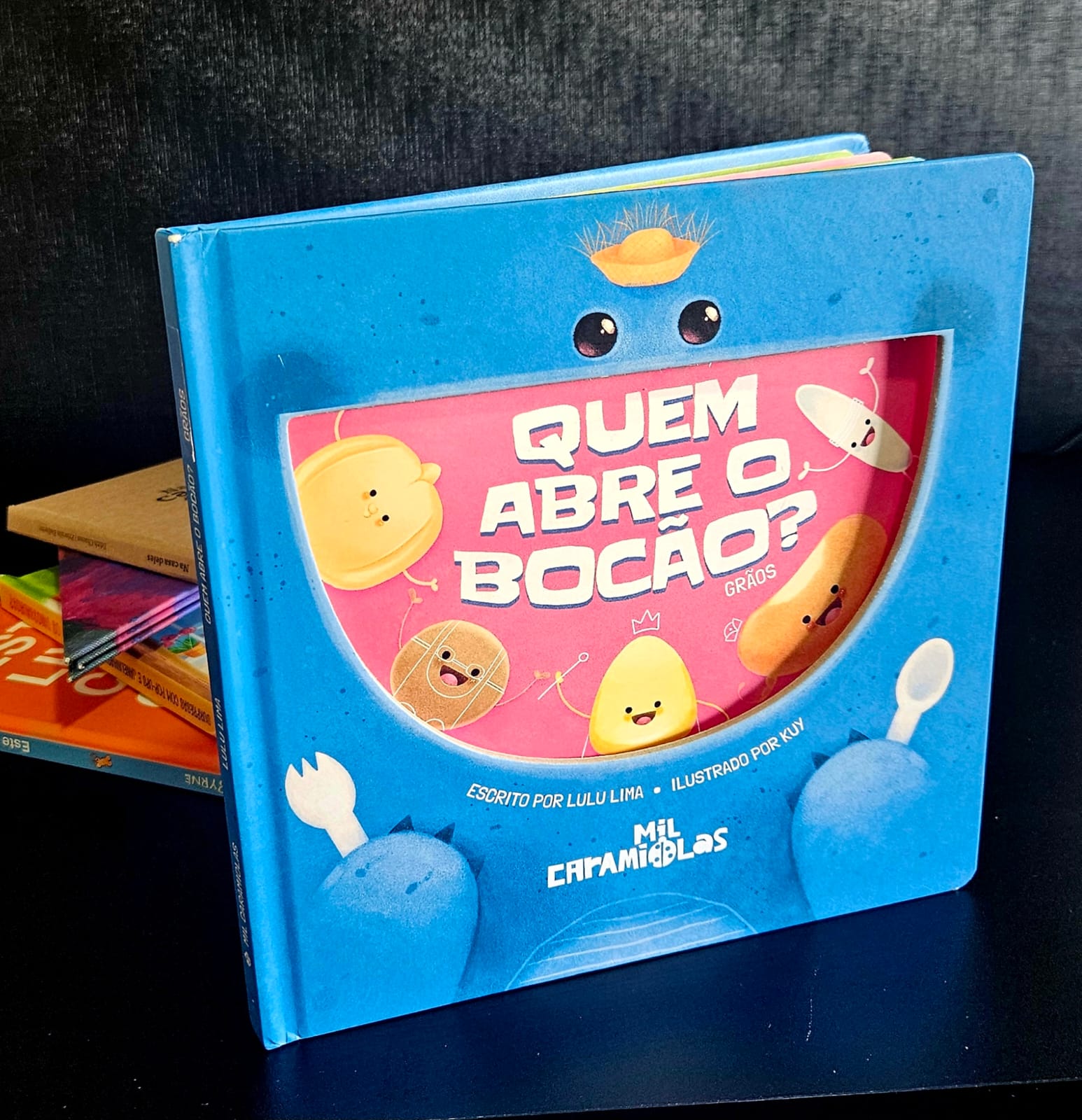 Foto colorida vertical de livro infantil e formato quadrado do livro "Quem abre o bocão". A obra tem na capa principal, em azul, tem dois olhinhos na parte de cima, e uma área vazada em formato de boca. Na página seguinte, visível atráves da área vazada, vários personagens, passarinhos e pintinhos, e uma superfície vermelha