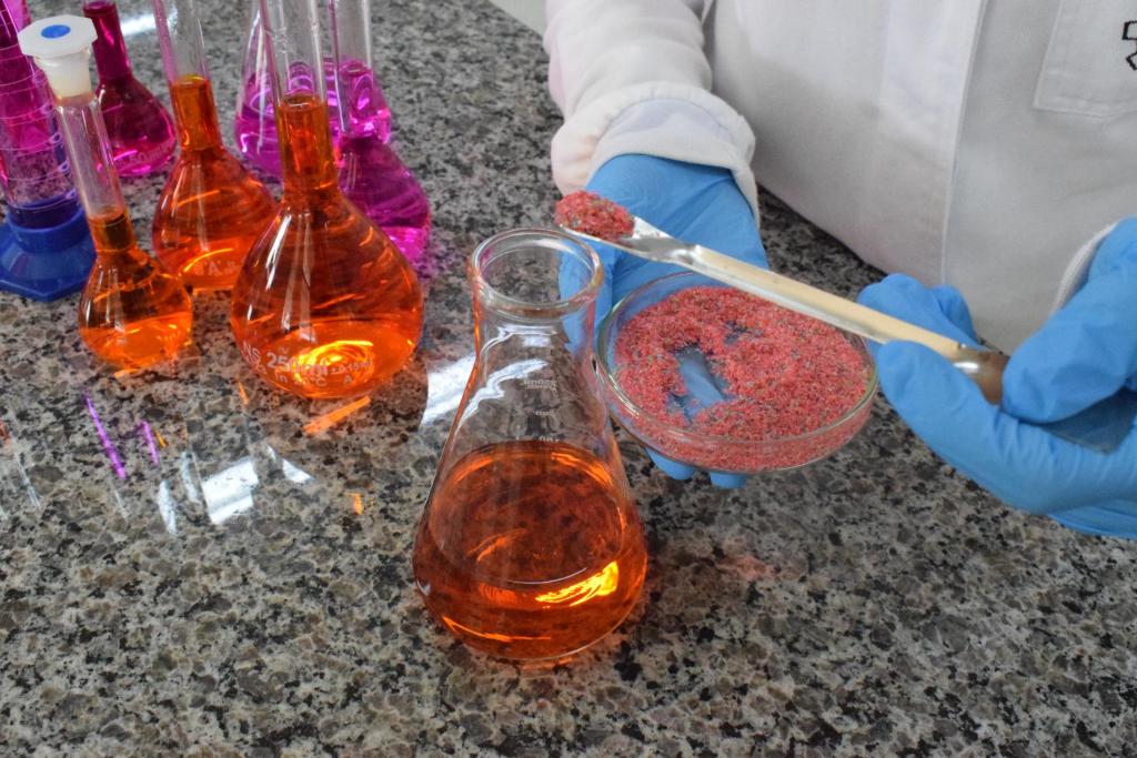 Foto colorida horizonal de bancada em laboratório de química. Uma pessoa com luvas azuis segura uma recipiente com pequenos pedaços de plástico e um ferramenta semelhante a uma pinça e coloc em um becker que contém um líquido cor alaranjada e translúcido. Ainda na bancada estão outras vidrarias com o mesmo líquido alarjado.
