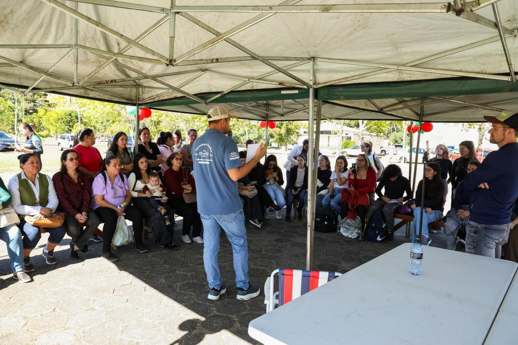Imagem horizontal colorida de uma roda de conversa realizada sob tendas da PoliFeira. Ao centro, um homem em pé fala ao público, enquanto dezenas de pessoas sentadas e em pé acompanham a atividade.