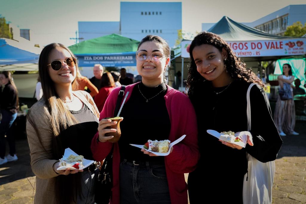 Imagem horizontal colorida de três jovens sorrindo em frente às bancas da PoliFeira, segurando fatias de bolo e uma cuia de chimarrão durante a celebração.