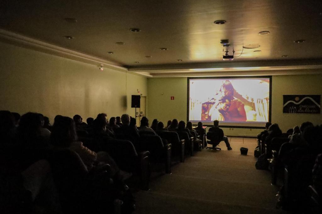 Foto colorida horizontal mostra uma sala escura de projeção com fileiras de cadeiras ocupadas por diversas pessoas sentadas, vistas de costas. Ao fundo, uma grande tela exibe uma cena do filme “Carandiru” com duas pessoas em destaque. O ambiente tem iluminação baixa, com luz principal vindo da projeção.