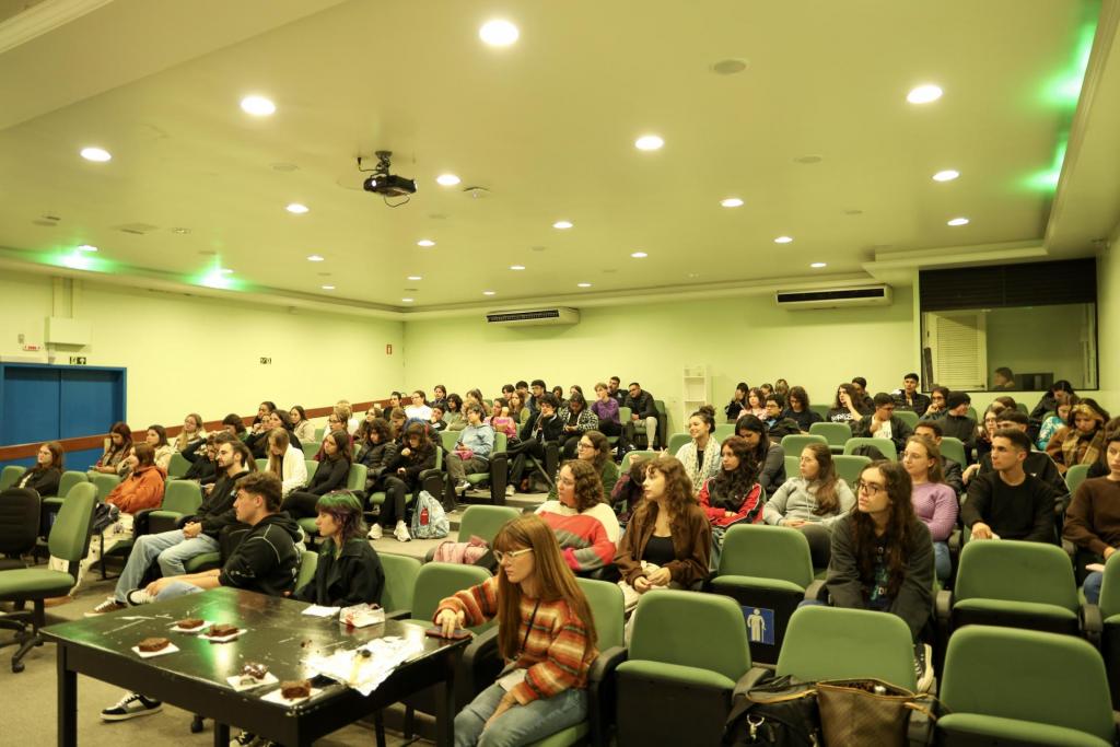 Foto colorida horizontal mostra uma sala ampla com várias fileiras de cadeiras verdes ocupadas por um público composto majoritariamente por jovens. As pessoas estão sentadas, assistindo a fala dos membros do projeto. O ambiente é bem iluminado.