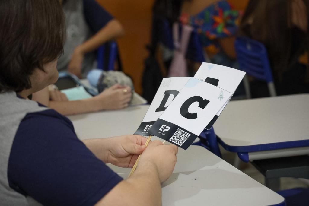 Sentada a uma mesa escolar, uma aluna segura placas de papel com as respostas para o quiz, as quais são representadas por letras (A, B, C e D). Ela veste uma camiseta predominantemente cinza, com mangas azul-marinho, tem pele branca e cabelo castanho e liso, de comprimento médio.