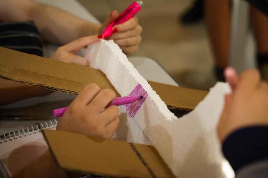 A foto ilustra a construção de um pequeno planador. A “fuselagem” é feita de isopor, enquanto que as asas são de papelão. Com canetinhas e canetar marca-texto, os alunos pintam o planador de cores como roxo e rosa.
