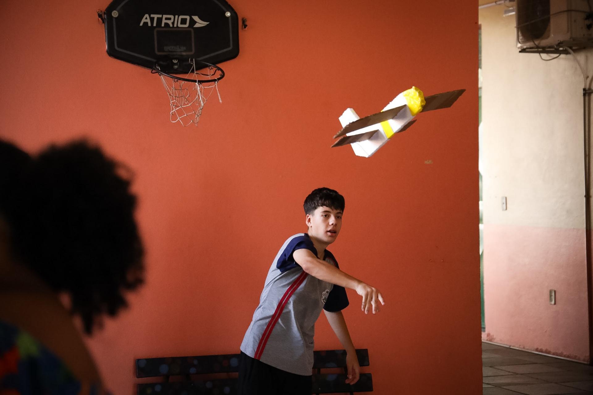 Tendo ao fundo uma parede de cor laranja (com uma cesta de basquete pendurada na parte de cima), um aluno da escola lança no ar um planador. O aluno veste uma camisa cinza; é moreno e tem cabelo curto e escuro.