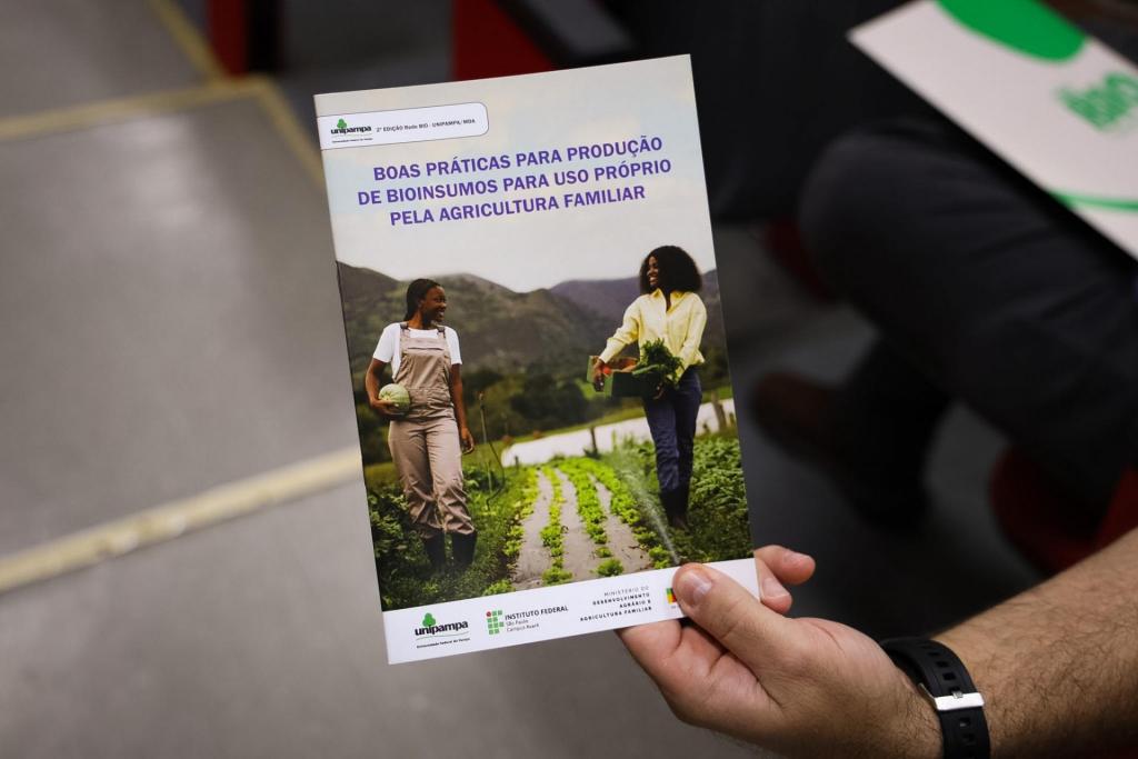 Foto colorida vertical. Uma pessoa segura uma cartilha sobre produção de bioinsumos para agricultura familiar. À esquerda aparece parte do corredor do auditório. À direita há poltronas e materiais do evento encobertos pela cartilha.