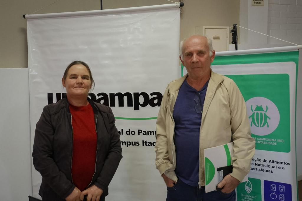 Foto colorida horizontal. Uma agricultora e um agricultor, que posam para a câmera durante evento da Rede Bio. À esquerda está um banner da Unipampa. À direita há um painel verde da Rede Bio. Ambos usam roupas casuais.