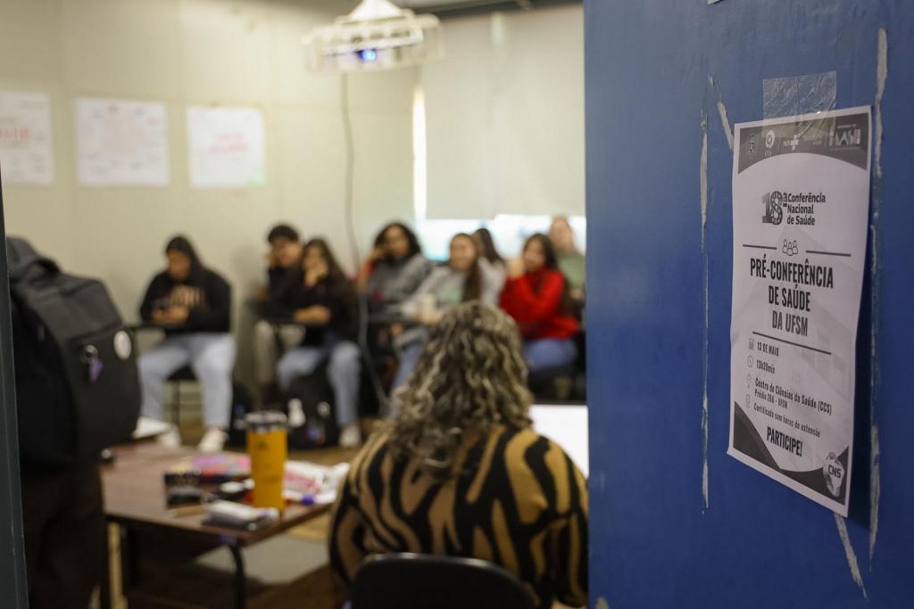 Foto colorida horizontal de grupo de pessoas sentadas em uma sala. A imagem mostra ainda uma porta com cartaz da conferência