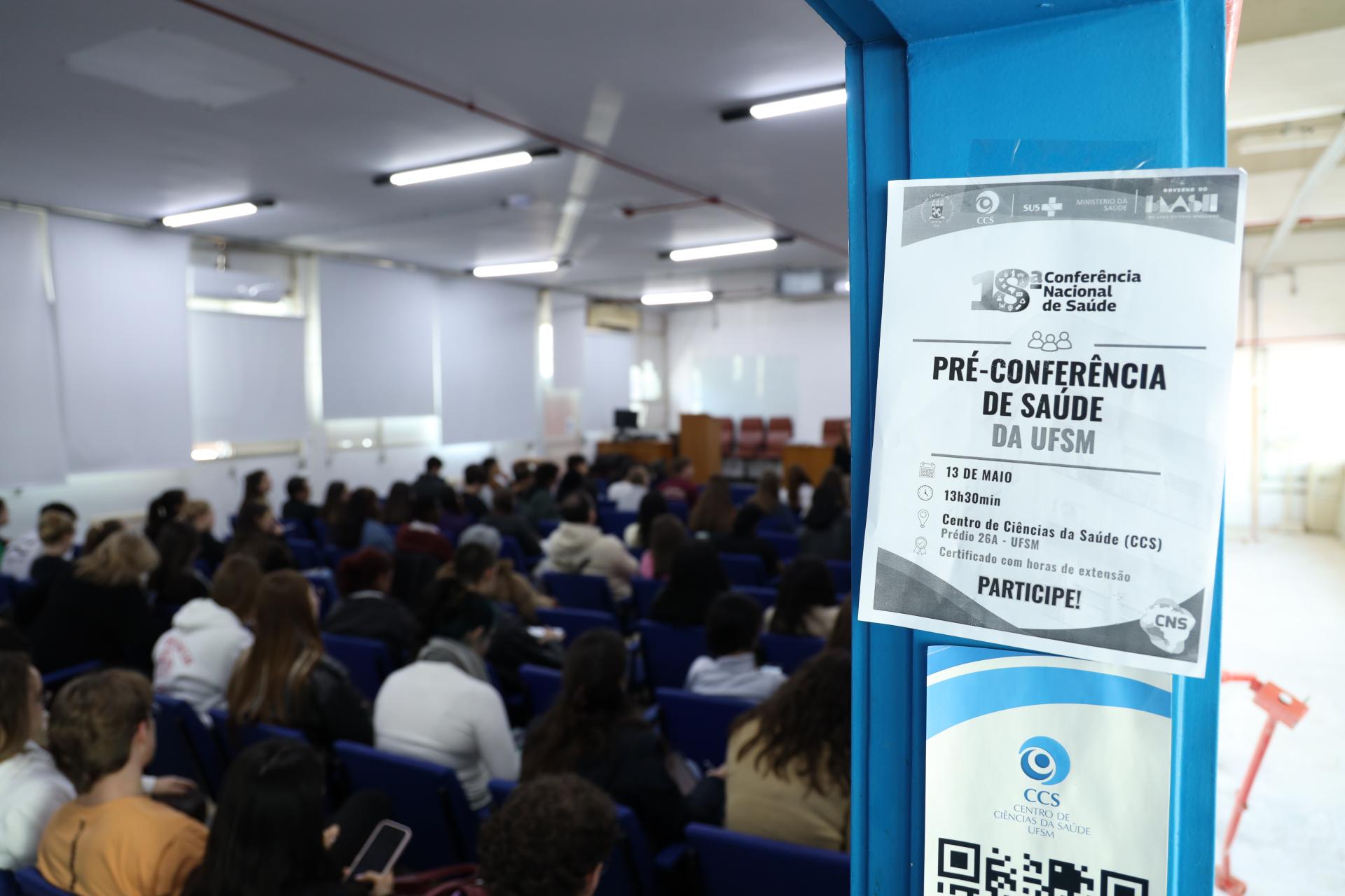 Foto colorida horizontal da pré-conferência de saúde. Em primeiro plano, cartaz fixado na porta com a indicação da conferência. Em segundo plano, o auditório lotado de participantes sentados nas cadeiras.