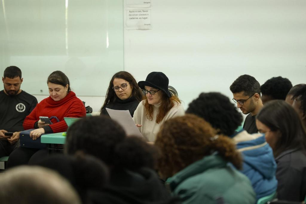Foto colorida horizontal de um grupo de pessoas sentadas em formato de círculo. Entre os participantes, está uma mulher com chapéu preto de tricô que segura um papel e faz a leitura. Ela é a professora Laís, a coordenadora da pré-conferência