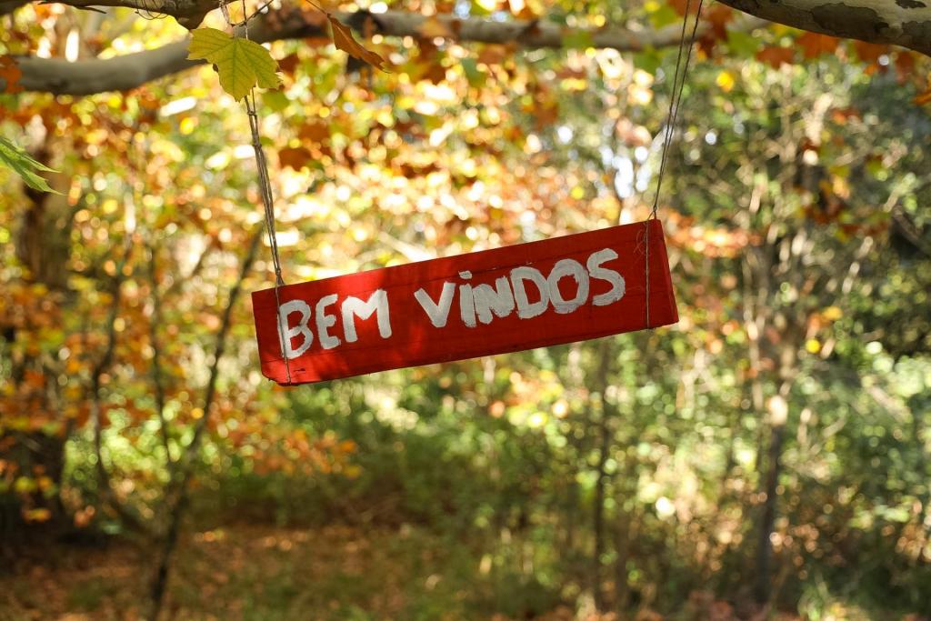 Foto colorida na horizontal mostra, ao centro, uma placa retangular vermelha com a inscrição “Bem Vindos” em letras brancas. A placa está pendurada por cordas em um galho de árvore que atravessa a parte superior da foto. Ao fundo, aparece uma área arborizada com folhas em tons de verde, amarelo e alaranjado, iluminadas pela luz do sol. A mistura de cores nas árvores remete à transição entre o verão e o outono, criando um cenário natural e acolhedor.