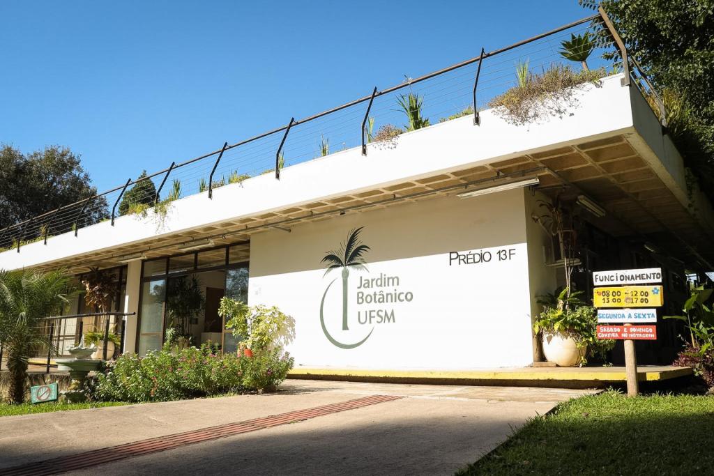 Foto colorida na horizontal mostra a fachada do prédio do Jardim Botânico da UFSM em um dia de céu limpo. O prédio branca ocupa grande parte da imagem. No centro da parede da frente, está o logotipo do Jardim Botânico UFSM, acompanhado do escrito “Prédio 13 F” em letras pretas à direita. Na parte superior do edifício há o telhado verde com plantas e grades metálicas de proteção. Em frente ao prédio, uma rampa de acesso de concreto em direção à entrada principal. Ao lado da rampa há canteiros com flores pequenas, além de vasos com plantas. À direita da imagem, próximo à entrada, há uma placa colorida indicando os horários de funcionamento do Jardim Botânico.