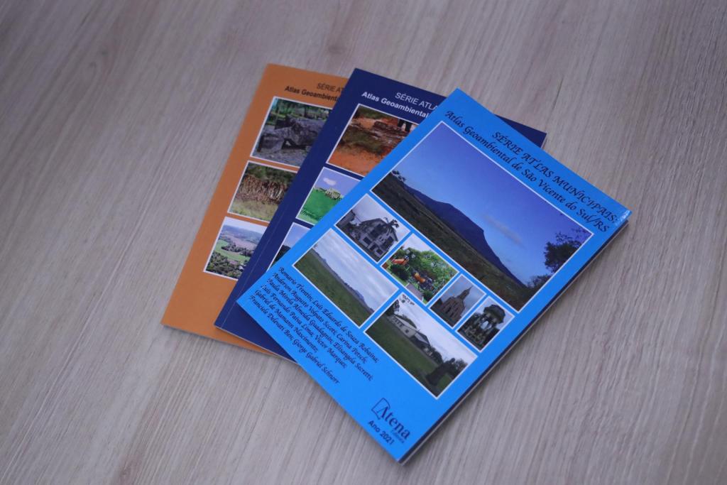 Foto horizontal colorida de três atlas geoambientais sob uma mesa de fundo claro. Os atlas estão fechados e têm capas com fotos de acidentes geográficos e fundos em azul claro, azul escuro e laranja